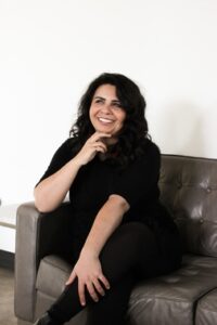 Author photo of Nisha J. Tuli, sitting on a leather couch and smiling, wearing a black top